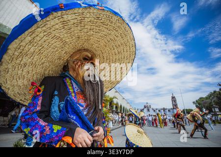 Flaches Porträt einer Folklore-Tänzerin, die eine alte Männermaske und einen riesigen Strohhut auf einem mexikanischen Stadtplatz trägt. Stockfoto
