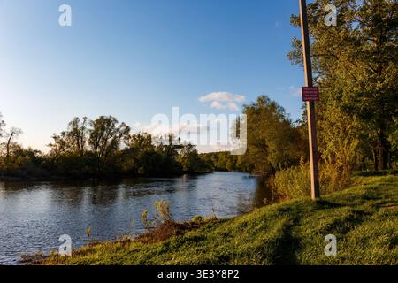 Ein ruhiger Fluss fließt an üppigen grünen Ufern unter einem riesigen blauen Himmel vorbei. Golden Hour Licht taucht das ruhige Wasser und das lebendige Laub auf, perfekt für einen Frieden Stockfoto