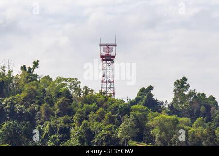 Ein rot-weißer Luftfahrradarturm, der sich auf einem üppig grünen, bewaldeten Hügel vor einem bewölkten Himmel befindet. Stockfoto