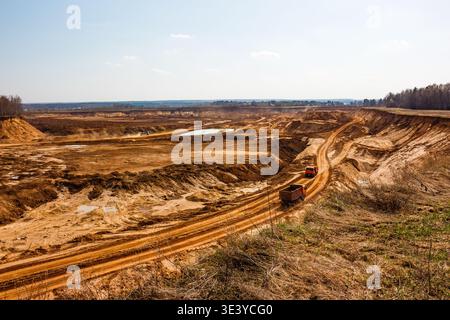 Riesige Sandmine im Tagebau mit schweren Lkw, die unter einem hellen Frühlingshimmel unbefestigte Straßen durchqueren und massive Erdbewegungen zeigen Stockfoto