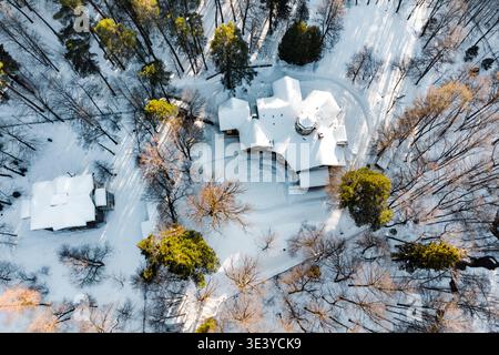 Die Drohne aus der Luft fängt ein schneebedecktes Luxushaus in einem dichten Winterwald ein, wo das Sonnenlicht lange Schatten über die weiße Landschaft wirft Stockfoto