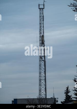 Der hohe Metallturm steht vor bewölktem Himmel. Der tragende Rahmen ragt über Dächern und Bäumen. Metallgitter für Geräte und Antennen. Leise Stockfoto
