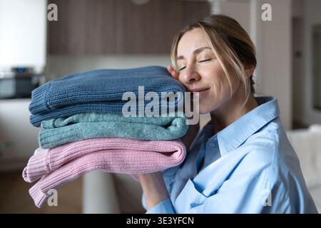 Zufriedene Frau mittleren Alters, die mit einem Lächeln saubere Aromawäsche hält und weiche Pullover zu Hause genießt Stockfoto
