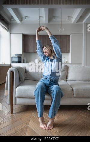 Zufriedene reife Frau, die die Arme nach oben streckt, während sie zu Hause auf einem gemütlichen Sofa sitzt und vor Freude lächelt. Stockfoto
