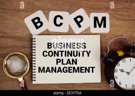 BCPM Business Continuity Plan Management Text auf Notizblock mit Lupe und Wecker auf hölzernem Hintergrund Stockfoto