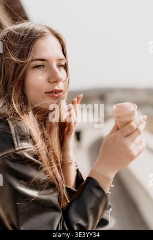 Frau Eiscreme draußen genießt süßes rosa Dessert in Waffelkegel während des sonnigen Tages in trendiger Lederjacke Stockfoto