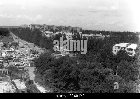 Ein historisches Schwarzweiß-Foto, das einen weiten Blick auf das Wohngebiet bietet. Ein geschäftiger Straßenmarkt mit zahlreichen Verkäufern, kleinen Zelten und einheimischen Einkäufern erstreckt sich entlang der belebten Straße. Im unmittelbaren Vordergrund befindet sich ein unvollendetes Baufundament inmitten von wildem Gras. Lange, mehrstöckige Wohnblöcke säumen den fernen Horizont. Dieser dynamische Schnappschuss aus dem Archiv erfasst wunderbar den postsowjetischen Straßenhandel und den friedlichen Donbass. Stockfoto