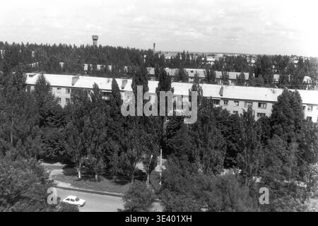 Sloviansk, Mitte der 1990er Jahre, Artem Microdistrikt (Lesnoy). Ein historisches Schwarzweiß-Foto, das eine erhöhte Perspektive auf eine ruhige Wohngegend bietet, die stark von Sommergrün umgeben ist. Ein klassischer fünfstöckiger Wohnblock ist teilweise hinter einer beeindruckenden Reihe von Pappelbäumen versteckt, die das Viertel säumen. Ein Hauswagen fährt leise entlang der Straße, während ein weiter entfernter Wasserturm die Skyline markiert. Dieses eindrucksvolle dokumentarische Bild bewahrt authentisch bürgerliches Design und Leben im friedlichen Donbass. Stockfoto