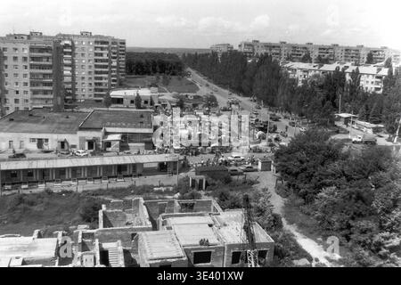 Sloviansk, Mitte der 1990er Jahre, Artem Microdistrikt (Lesnoy). Ein historisches Schwarzweiß-Foto mit einem weitläufigen Blick auf die geschäftige Wohngegend. Im Vordergrund steht ein unvollendetes Betonfundament inmitten von wildem Grün. Weiter unten gibt es einen lebhaften, spontanen Straßenmarkt mit kleinen Verkaufsständen und geschäftigen Menschenmassen entlang der Hauptstraße. Riesige mehrstöckige Blöcke runden die ferne Skyline ab. Dieser detailreiche Dokumentarrahmen bewahrt die postsowjetische urbane Mikroökonomie und den friedlichen Donbass akribisch. Stockfoto