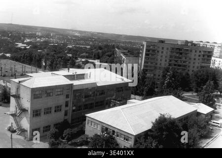 Sloviansk, Mitte der 1990er Jahre, Artem Microdistrikt (Bäckerei-Bereich). Ein historisches Schwarzweiß-Foto, das eine weitläufige erhöhte Perspektive über eine gemischte Stadtzone einfängt. Im Mittelpunkt steht ein großes, mehrgeschossiges Industriebäckereigebäude mit flachem Dach und externen Brandausgängen. Hinter den üppigen Sommerbäumen erstrecken sich hoch aufragende Wohnblocks durch die städtische Landschaft. Dieser authentische Schnappschuss aus dem Archiv zeigt wunderbar die postsowjetische zivile Infrastruktur, industrielle Architektur und das tägliche Leben im friedlichen Donbass. Stockfoto