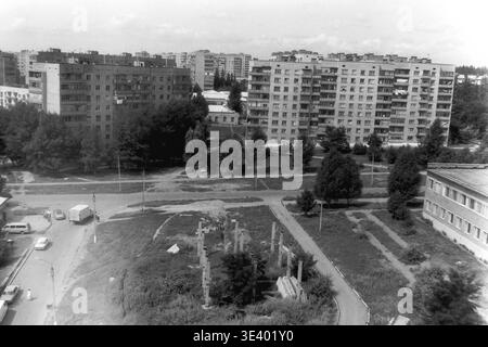 Sloviansk, Mitte der 1990er Jahre, Artem Microdistrikt (Bäckerei-Bereich). Ein historisches Schwarzweiß-Foto, das einen erhöhten Blick auf ein sich entwickelndes Wohnviertel zeigt. Im Vordergrund erheben sich Betonsäulen einer verlassenen Baustelle aus dem bewachsenen Gras. Ein weißer Nutzfahrzeug und ein paar Fußgänger befahren die asphaltierte Straße. Massive mehrstöckige Paneelgebäude stehen hinter dicken Sommerbäumen. Dieses realistische dokumentarische Bild stellt die postsowjetische Stadterweiterung und das Leben im friedlichen Donbass authentisch dar. Stockfoto