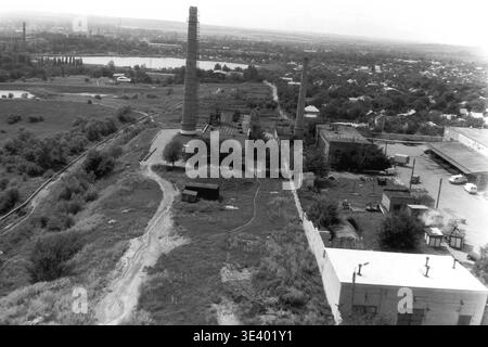 Sloviansk, Mitte der 1990er Jahre, Artem Microdistrikt (Bäckerei-Bereich). Ein historisches Schwarzweiß-Foto mit atemberaubendem Blick über die lokalen Industrieanlagen. Zwei hoch aufragende Backsteinkamine der Bäckerei dominieren das Zentrum, umgeben von verschiedenen Versorgungsgebäuden und gepflasterten Höfen. Hinter dem dichten Sommergrün erstrecken sich ein ruhiger See und weitläufige Wohngebiete zum Horizont hin. Dieser stimmungsvolle Dokumentarrahmen bewahrt die postsowjetische industrielle Ökologie und das Leben im friedlichen Donbass. Stockfoto