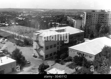 Sloviansk, Mitte der 1990er Jahre, Artem Microdistrikt (Bäckerei-Bereich). Ein historisches Schwarzweiß-Straßenfoto, das ein weites, erhöhtes Panorama eines wichtigen kommunalen Unternehmens bietet. Das stabile Flachdach des örtlichen Bäckereibetriebs nimmt den Vordergrund ein, flankiert von geparkten Gewerbewagen auf der linken Seite. Üppige, ältere Bäume verbergen teilweise die hohen Wohnblöcke, die im fernen Hintergrund thronen. Dieses herausragende Archivbild dokumentiert makellos das postsowjetische industrielle Erbe und den Rhythmus des friedlichen Donbass. Stockfoto