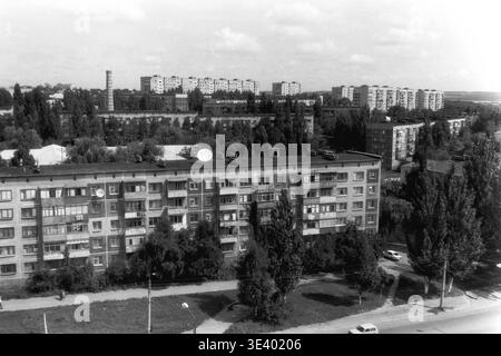 Sloviansk, Mitte der 1990er Jahre, Artem Microdistrikt (Molodiozhnaya-Gebiet). Ein historisches Schwarzweiß-Foto, das sich auf ein klassisches, langes fünfstöckiges Wohngebäude mit Paneelverkleidung von oben konzentriert. Interessanterweise sind mehrere frühe Satellitenschüsseln auf dem flachen Dach montiert. Dicke Sommerbäume umgeben die Architektur stark, während ein einzelnes weißes Hausfahrzeug die gepflasterte Straße in der unteren rechten Ecke entlang fährt. Dieses unglaublich authentische Archivbild dokumentiert die postsowjetische Wohninfrastruktur und ruhige Routinen im friedlichen Donbass. Stockfoto