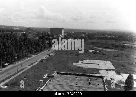 Sloviansk, Mitte der 1990er Jahre, Artem Microdistrikt (Molodiozhnaya-Gebiet). Ein historisches Schwarzweiß-Foto mit einem weiten Hochwinkel über einem sich entwickelnden Stadtrand. Auf der breiten Asphaltstraße fahren einige weit entfernte Autos entlang einer dichten Gasse mit hohen Sommerbäumen. Auf der rechten Seite befindet sich ein weitläufiges, verlassenes Betonfundament, das sich bis zum Horizont erstreckt. Dieser majestätische Dokumentarrahmen fängt die postsowjetische kommunale Expansion und die Atmosphäre des friedlichen Donbas ein. Stockfoto