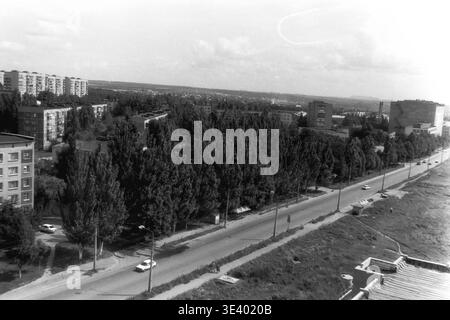 Sloviansk, Mitte der 1990er Jahre, Artem Microdistrikt (Molodiozhnaya-Gebiet). Ein historisches Schwarzweiß-Foto, das eine wunderschöne, erhöhte Perspektive entlang einer markanten, von Bäumen gesäumten Stadtallee einfängt. Ein weißer Pkw nähert sich der Kamera auf der breiten Asphaltstraße, flankiert von einer beeindruckenden, undurchdringlichen Mauer aus hohen Sommerpappeln auf der linken Seite. Die weitläufigen Wohnblöcke des Viertels sind im Hintergrund zu sehen. Diese eindrucksvolle Archivaufnahme bewahrt die postsowjetische urbane Mobilität und das regelmäßige Leben im friedlichen Donbass. Stockfoto