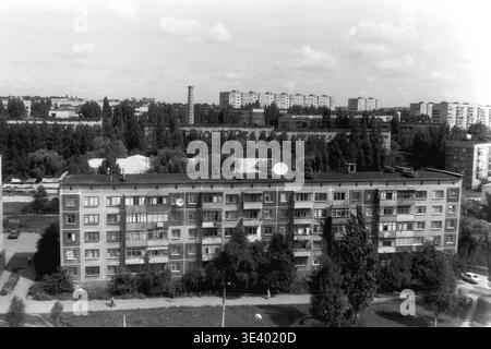 Sloviansk, Mitte der 1990er Jahre, Artem Microdistrikt (Molodiozhnaya-Gebiet). Ein historisches Schwarzweiß-Foto, das eine breite, erhöhte Perspektive auf ein klassisches Wohnviertel bietet. Ein langes fünfstöckiges Paneelgebäude befindet sich im Vordergrund, auf dem auf dem flachen Dach frühe Satellitenschüsseln angebracht sind. Die Einheimischen spazieren auf den gepflasterten Pfaden, umgeben von üppigen Sommerbäumen. Im fernen Hintergrund ist ein hoher Industrieschornstein zu sehen. Dieses detaillierte Archivbild dokumentiert perfekt postsowjetische Wohngebäude und das Leben im friedlichen Donbass. Stockfoto
