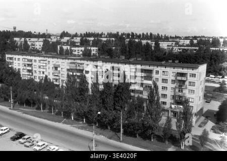 Sloviansk, Mitte der 1990er Jahre, Artem Microdistrikt (Molodiozhnaya-Gebiet). Ein historisches Schwarzweiß-Foto, das einen weitläufigen Hochwinkelblick über eine dicht besiedelte Stadtzone zeigt. Ein klassischer fünfstöckiger Wohnblock erstreckt sich über den Rahmen, der teilweise von einer dicken Linie hoher Pappeln verdeckt wird. Ein weißes Hausfahrzeug fährt links, während Fußgänger auf dem breiten Bürgersteig spazieren. Dieser fesselnde dokumentarische Schnappschuss bewahrt die postsowjetische städtische Infrastruktur und das tägliche Leben im friedlichen Donbass authentisch. Stockfoto