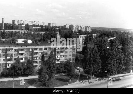 Sloviansk, Mitte der 1990er Jahre, Artem Microdistrikt (Molodiozhnaya-Gebiet). Ein historisches Schwarzweiß-Foto, das einen ruhigen Blick auf einen grünen Wohnsektor zeigt. Die Szene konzentriert sich auf einen mehrstöckigen Wohnblock mit mehreren Satellitenantennen auf dem Dach. Auf der rechten Seite fährt ein weißer Pkw die Asphaltstraße hinunter, während die Bewohner gemütlich im Schatten der dichten Sommerbäume spazieren gehen. Dieser realistische Archivrahmen veranschaulicht wunderbar die postsowjetische städtische Ökologie und das Leben im friedlichen Donbass. Stockfoto