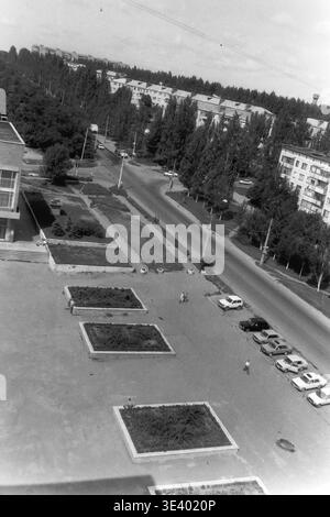 Sloviansk, Mitte der 1990er Jahre, Artem Microdistrikt (Bereich des Pädagogischen Instituts). Ein historisches Schwarzweiß-Foto mit beeindruckendem Blick über einen geräumigen Stadtplatz. Der gepflasterte Bereich verfügt über mehrere rechteckige Blumenbeete, mit einer Reihe verschiedener Wohnwagen, die ordentlich auf der rechten Seite parken. Einheimische werden bei einem gemütlichen Spaziergang über den offenen platz in Richtung der breiten, von Bäumen gesäumten Stadtallee beobachtet. Dieser außergewöhnlich detaillierte Archivfoto erfasst die postsowjetische Infrastruktur und das Leben im friedlichen Donbass. Stockfoto