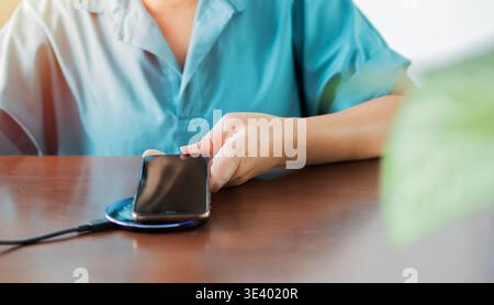 Weibliche Ladebatterie des Mobiltelefons mit drahtlosem Ladegerät im Tisch. Mobiltelefon in der Nähe eines kabellosen Ladegeräts, moderne Lifestyle-Technologie, versteckt Stockfoto