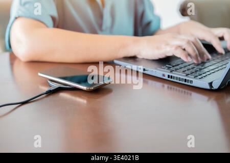 Smartphone mit kabellosem Ladegerät neben Frau mit Laptop am Arbeitsplatz. Smartphone wird auf einem Ladepad aufgeladen. Modernes Arbeitsplatzzubehör-Konzept. Stockfoto