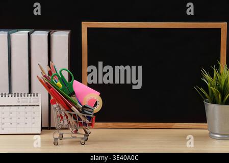 Schreibtisch im Klassenzimmer mit Tafel und Schreibwaren im Einkaufswagen Stockfoto