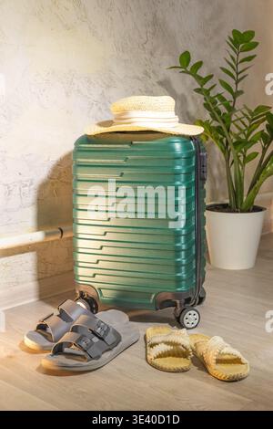 Stilvolle Komposition: Grüner Hartschalenkoffer mit strukturierten Rippen, Strohhut oben, grauen und beigefarbenen Hausschuhen vorne. Stockfoto