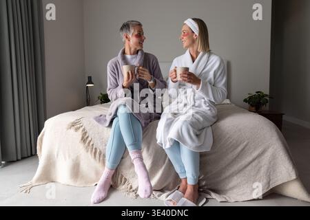 Ältere Mutter und Tochter sitzen auf dem Bett im Schlafzimmer, tragen Bademäntel und Augenflecken und schlürfen Tassen Stockfoto