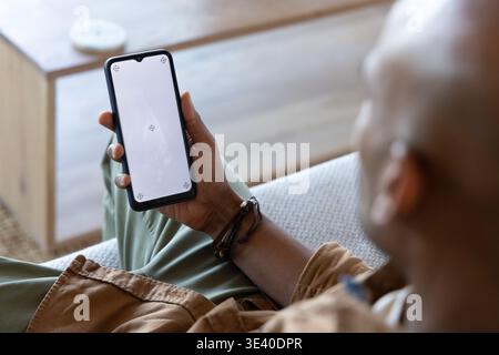 Afroamerikaner sitzt auf dem Sofa und hält Telefonmockup in braunem Hemd und Armbändern, Kopierraum Stockfoto