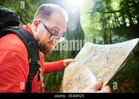 Nahaufnahme der bärtigen Touristenmannskarte, während sie vor einem großen Felsen in dichtem Wald steht. Reisende mit grauem Rucksack und Brille, planen Outdoor-Abenteuer, Wander- oder Kletterrouten. Stockfoto