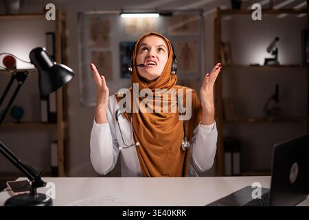 Verärgerte weibliche muslimische Ärztin im Hijab oder Arzttherapeutin im weißen Mantel Tragen Sie Headset mit Mikrofon arbeiten mit Laptop schreiende Hände nach oben während n Stockfoto