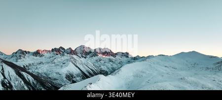 Ein atemberaubender Panoramablick auf die schneebedeckten Berggipfel unter einem leuchtenden rosa und blauen Sonnenuntergang. Die Szene fängt die heitere Schönheit der alpi ein Stockfoto