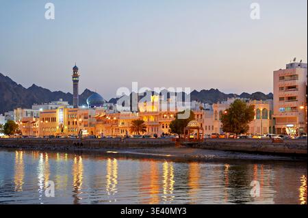 Der Sonnenuntergang über der Uferpromenade in Maskat Oman zeigt Gebäude und Berge. Die Lichter der Stadt spiegeln sich bei Einbruch der Nacht auf der Wasseroberfläche. Stockfoto