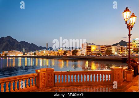 Der Sonnenuntergang strahlt einen Glanz über der Uferpromenade in Maskat Oman. Lichter von Gebäuden spiegeln sich im ruhigen Wasser und schaffen eine lebendige Atmosphäre. Stockfoto