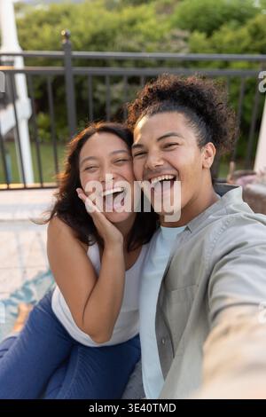 Diverse Paare lachen und machen Selfie auf gefliestem Balkon mit Smartphone, Frau im weißen Tank. Terrasse, Paar, draußen, Telefon, offen, lässig, Tageslicht || Model veröffentlicht Stockfoto