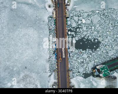 Aus der Vogelperspektive einer Brücke, die durch ein Mosaik aus eisigem Blau und weiß schneidet, mit einem Lastkahn, der durch die zerrissenen Gewässer von Danzig, Polen, fährt. Stockfoto