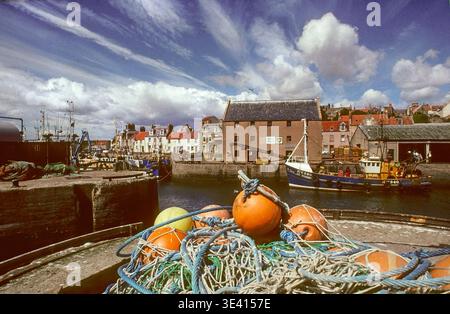 Am Kai im Hafen von Pittenweem befinden sich unzählige Angelausrüstung, leuchtend orange Bojen und gewickelte Seile mit Trawlern, Steinbauten und dramatischen Wolken Stockfoto