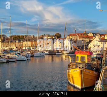 Der Hafen von Anstruther leuchtet in warmem goldenem Licht, während farbenfrohe Fischerboote neben einem Steinufer mit Geschäften am Ufer, Anlegejachten und traditionellen Stockfoto