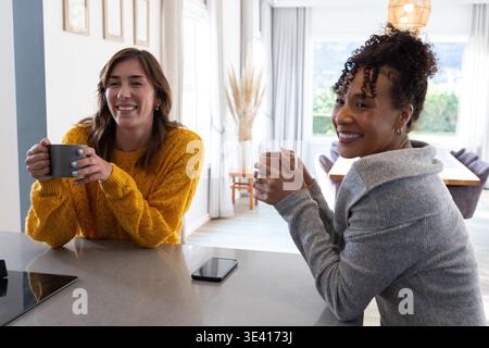 Verschiedene weibliche Freunde sitzen auf der Kücheninsel in Pullover mit Tassen und lächelnden Telefonen in der Nähe Stockfoto