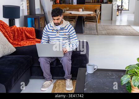 Indischer Mann mit gestreiftem Hoodie, der auf silbernem Laptop auf dunklem Sofa im Wohnzimmer arbeitet Stockfoto