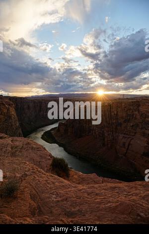 Der sich windende Fluss fließt durch die tiefe Sandsteinschlucht, während die Sonne am Horizont unter dramatischen Wolken untergeht, die von der erhöhten Klippe aus betrachtet werden Stockfoto