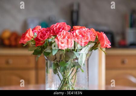 Ein Blumenstrauß aus rosa Rosen in Korallenrosen, elegant in einer klaren Glasvase angeordnet, auf einem Holztisch in gemütlicher Atmosphäre platziert. Das weiche Tageslicht. Stockfoto