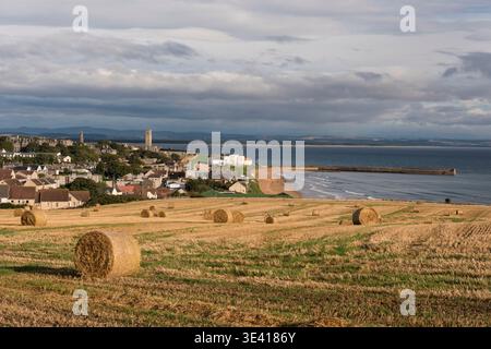 Ein Panoramablick über erntete Felder mit runden Heuballen in Richtung St Andrews Stadt, Sandstrand und Pier an der Fife Küste, sanftes Licht und dramatische cl Stockfoto