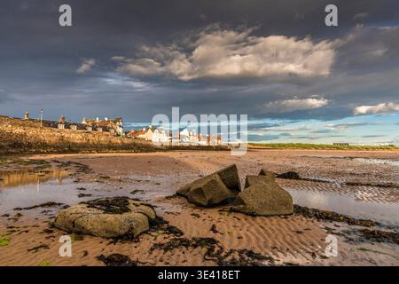 Elie Beach bei Ebbe zeigt felsige Betonblöcke, welligen Sand und ein historisches Küstendorf unter dramatischen Sturmwolken, das an Einsamkeit und Küste erinnert Stockfoto