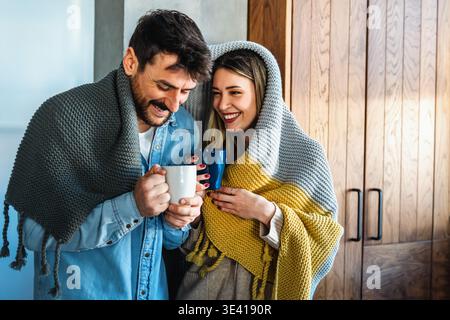 Fröhliches, junges Paar, bedeckt mit einer Decke, die sich umschließt, während man zu Hause Tassen Tee hält Stockfoto