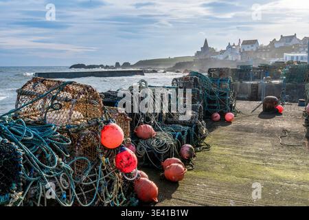 Fischernetze, Hummertöpfe und helle Bojen stapeln sich auf einem verwitterten Hafenpier von St. Monans mit Blick auf die Nordsee und ein malerisches Küstendorf Stockfoto