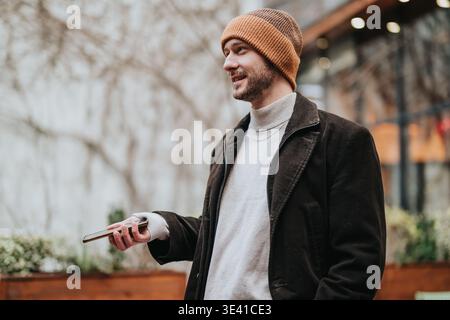 Junger Mann in Mütze und Mantel lächelt draußen, während er ein Smartphone hält Stockfoto