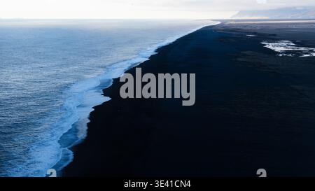 Blick aus der Vogelperspektive auf den Schwarzen Sandstrand von Reynisfjara aus Dyrhólaey, Südisland, mit Wellen des Atlantiks und fernen Küstenbergen. Stockfoto