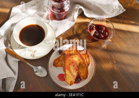Französischer Toast mit Kirschmarmelade und einer Tasse schwarzen Kaffee auf Holztisch. Goldene Brioche-Scheiben mit Beerensirup und Espresso für ein gemütliches Frühstück Stockfoto